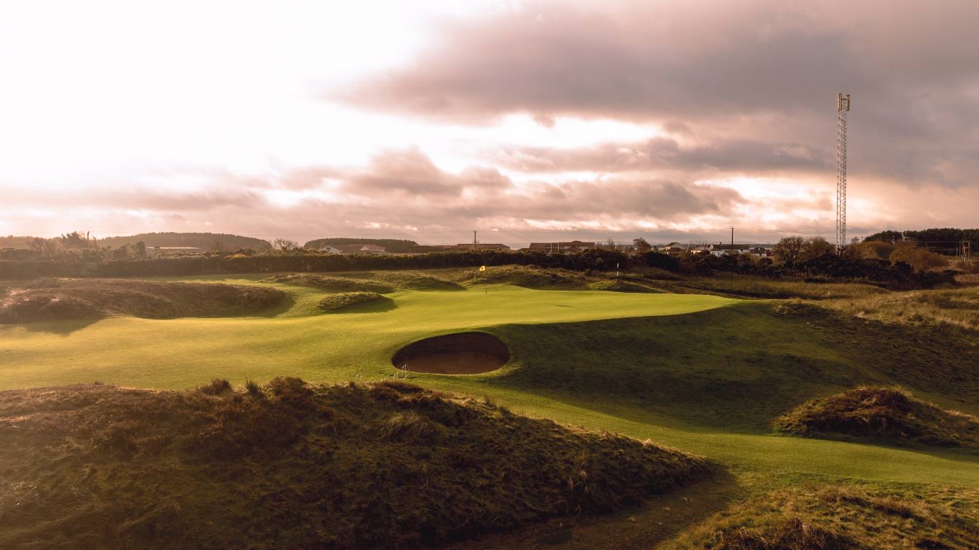 The sixth green at Panmure in evening light, the pot bunker short and right of the putting surface with heather in the foreground